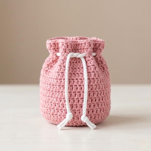 Puede incluir: Una pequeña bolsa rosa con cordón de ganchillo. La bolsa está hecha de un hilo texturizado y presenta un cierre de cordón blanco. La bolsa es redonda y tiene una estética sencilla y hecha a mano. El fondo es de color neutro.