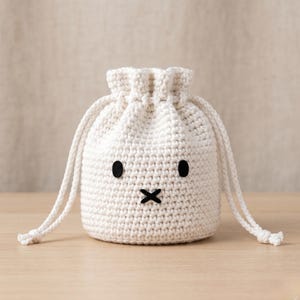 May include: A white crochet drawstring bag with a cartoon face. The bag features black oval eyes and a black X-shaped mouth. The bag is sitting on a wooden surface.