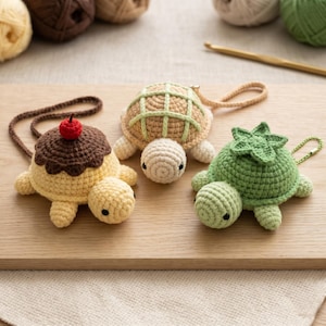 May include: Three crocheted turtle keychains in yellow, tan, and green, each with unique shell designs. One resembles a sundae with a cherry, another has a grid pattern, and the third has a leaf-like design. They are displayed on a wooden surface.