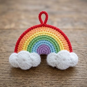 Könnte beinhalten: Ein gehäkeltes Regenbogen-Ornament mit konzentrischen Bögen aus rotem, orangem, gelbem, grünem, blauem und violettem Garn. Der Regenbogen wird an der Basis von zwei weißen gehäkelten Wolken verankert und hat oben eine kleine rote Garnschlaufe.