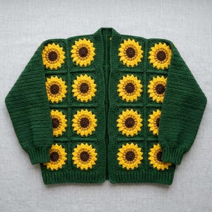 Puede incluir: Un cárdigan de ganchillo de manga larga con una base de color verde oscuro y un patrón repetitivo de girasoles amarillos con centros marrones. La prenda está construida con motivos cuadrados, cada uno con un girasol, y mangas verdes lisas.