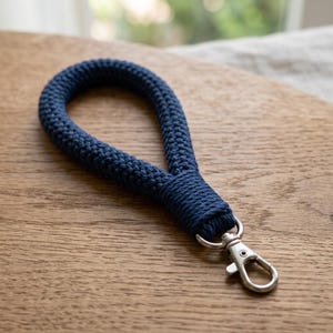 May include: A dark navy blue wrist lanyard crafted from braided cord in a continuous loop. The cord is tightly wound at the base where it connects to a silver-toned metal lobster clasp, providing a secure attachment point for keys or accessories.