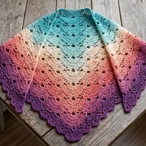 Pode incluir: Um xale triangular de crochê com um padrão de cores degradê que transita do azul-petróleo no centro superior para creme, coral e, finalmente, roxo profundo nas bordas festonadas. A peça é feita com um intrincado design de pontos em leque.