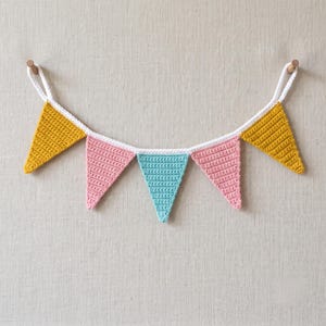 Puede incluir: Una guirnalda de banderines de ganchillo con cinco banderas triangulares en amarillo mostaza, rosa suave y azul turquesa. Las banderas están unidas por un cordón blanco de ganchillo, con bucles en cada extremo para colgar la decoración.
