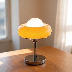 Könnte beinhalten: Eine Tischlampe im Retro-Stil mit einer hellen, kugelförmigen Lichtquelle. Die Lampe hat einen Chromschaft und -fuß, der einen breiten, orange-gelben Glasschirm trägt. Die Lampe steht auf einem Holztisch.
