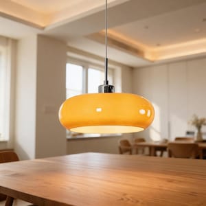 Könnte beinhalten: Eine moderne, orange-gelbe Pendelleuchte mit einem abgerundeten, länglichen Schirm. Das Licht hängt an einem dünnen, silbernen Kabel über einem Holzesstisch. Der Raum hat eine warme, einladende Atmosphäre.