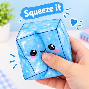 Könnte beinhalten: Ein hellblaues, würfelförmiges Squishy-Spielzeug mit einem Muster aus weißen Sternen und einem Cartoon-Gesicht mit schwarzen Augen, einem kleinen Lächeln und rosa Wangen. Über dem Würfel steht in einer blauen Sprechblase der Text Squeeze it.