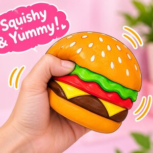 Puede incluir: Un juguete de hamburguesa blanda con un pan dorado con semillas de sésamo blancas, lechuga verde, una rodaja de tomate rojo, queso amarillo y una carne marrón. Un globo de texto rosa arriba contiene el texto blanco Squishy & Yummy!