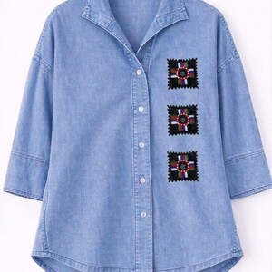 Puede incluir: Camisa vaquera azul claro con tres parches bordados. Cada parche es un cuadrado negro con un diseño floral colorido en el centro. La camisa tiene una botonadura frontal, un cuello y mangas tres cuartos.