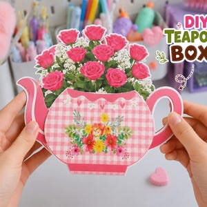 May include: A DIY teapot box featuring a pink and white gingham pattern with a floral bouquet of pink roses and white baby's breath. The teapot shape includes a central floral arrangement and small heart accents. The text DIY TEAPOT BOX is visible.