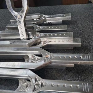 May include: A collection of silver metal tuning forks. Each fork features a unique design with geometric patterns and small holes along the sides. The forks are stacked on a dark grey surface.