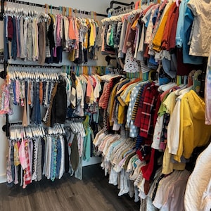 May include: A closet filled with children's clothing, including shirts, pants, and dresses, hanging on multiple racks. The clothes are in various colors and patterns, with a variety of hanger colors.