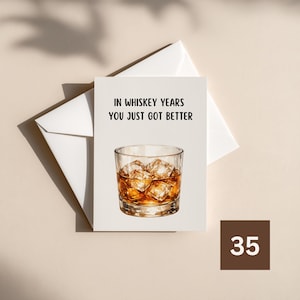 May include: A white greeting card featuring a centered illustration of a glass of amber-colored whiskey with ice cubes. The text printed in black above the glass reads, IN WHISKEY YEARS YOU JUST GOT BETTER. A white envelope is positioned behind the card.
