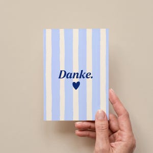 May include: A rectangular greeting card featuring a vertical striped pattern in light blue and cream. The center of the card displays the word "Danke." in a dark blue serif font, positioned directly above a small, solid dark blue heart icon.