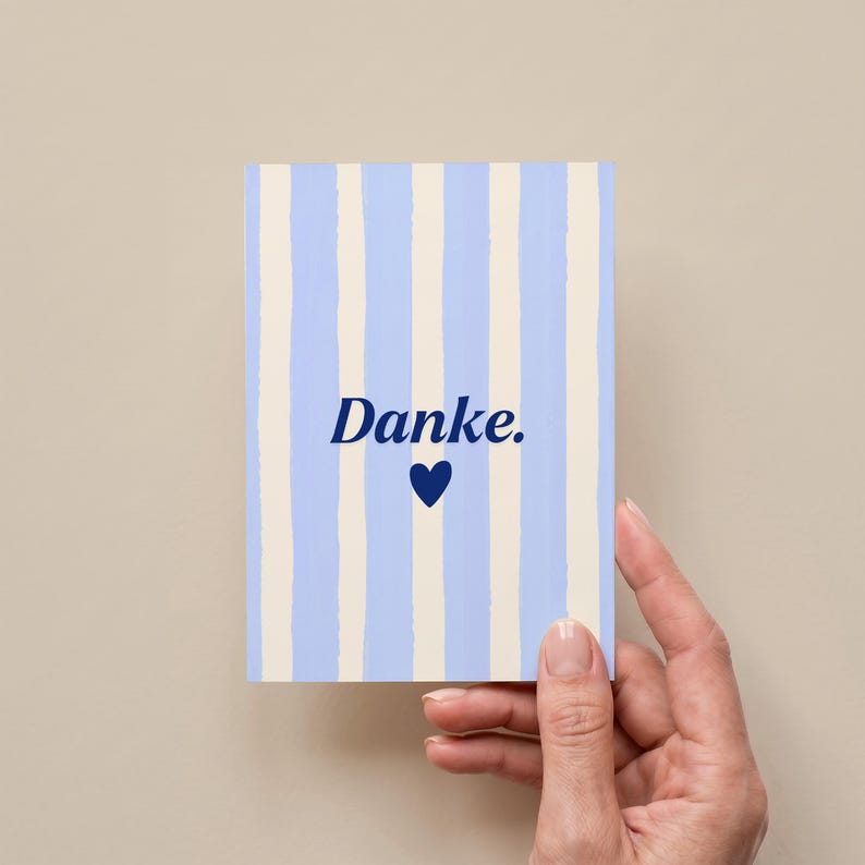May include: A rectangular greeting card featuring a vertical striped pattern in light blue and cream. The center of the card displays the word "Danke." in a dark blue serif font, positioned directly above a small, solid dark blue heart icon.