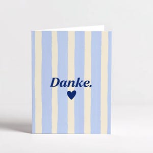 May include: A folded greeting card featuring a pattern of vertical light blue and cream stripes. The front of the card displays the word "Danke." in dark blue text, positioned directly above a small, solid dark blue heart icon.