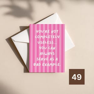 Puede incluir: Una tarjeta de felicitación rectangular con un patrón de rayas verticales en rosa y rosa claro. El texto en blanco dice: YOU'RE NOT COMPLETELY USELESS... YOU CAN ALWAYS SERVE AS A BAD EXAMPLE. La tarjeta está sobre un sobre blanco.