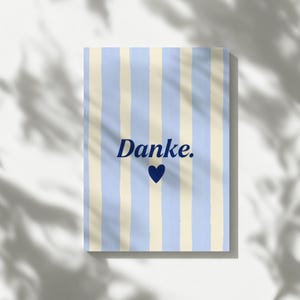 May include: A rectangular card featuring a vertical striped pattern in light blue and cream. Centered on the card is the word "Danke." written in a dark blue serif font, positioned directly above a solid dark blue heart icon.
