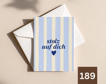 May include: A rectangular greeting card featuring vertical light blue and cream stripes with the dark blue text stolz auf dich and a small dark blue heart icon centred on the front, accompanied by a plain white envelope tucked behind the card.