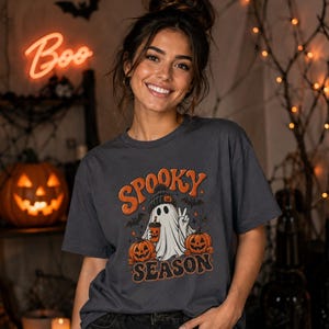 Può includere: Una t-shirt grigio antracite con la grafica di un fantasma che indossa un berretto, tiene in mano una tazza di caffè e fa il segno di vittoria. Il fantasma è affiancato da due zucche e pipistrelli, con il testo arancione "SPOOKY SEASON" sopra e sotto.