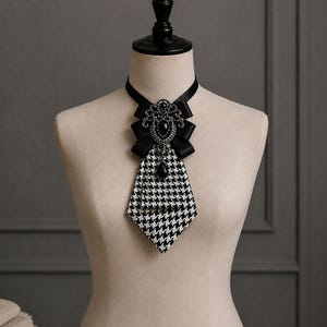 May include: A black and white houndstooth patterned necktie accessory featuring a black satin ribbon collar. The center is adorned with a decorative black brooch embellished with clear crystals and a central black teardrop-shaped pendant.