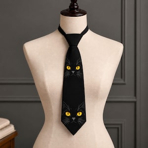 Könnte beinhalten: Eine schwarze Krawatte mit einem wiederkehrenden Grafikdruck eines schwarzen Katzengesichts mit leuchtend gelben Augen und weißen Schnurrhaardetails. Die Krawatte wird auf einem neutralfarbenen Schaufensterpuppen-Torso präsentiert.
