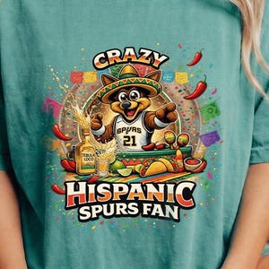 May include: Teal t-shirt with a graphic of a cartoon fox in a sombrero and Spurs jersey, holding tequila. Text reads "CRAZY HISPANIC SPURS FAN". The design incorporates chilli peppers and festive decorations.