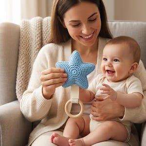 May include: A light blue star-shaped crochet rattle featuring a smooth, round wooden ring handle attached by a cream-colored fabric strap. The star is textured with visible yarn stitches, providing a soft, tactile surface for grasping and play.