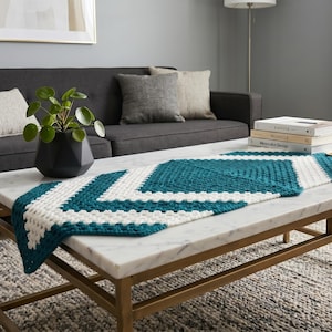 May include: A rectangular crochet table runner featuring a geometric chevron pattern in alternating bands of deep teal and off-white yarn. The textile has a textured, knotted stitch construction and is draped lengthwise across a white marble surface.