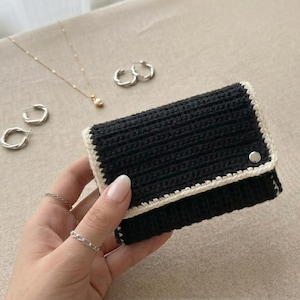 May include: A rectangular black crochet wallet featuring a cream-colored border and a single silver-toned metal snap closure. The wallet is constructed with a horizontal ribbed stitch pattern and is finished with a clean, contrasting edge trim.