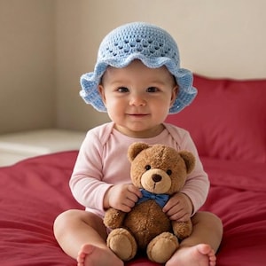 May include: A baby wearing a light blue crochet hat and a pink onesie sits on a red bed, holding a brown teddy bear with a blue bow tie. The baby is smiling, and the background includes a window and red bedding.