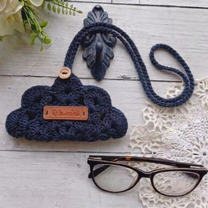 May include: Navy blue crocheted glasses case with a matching cord, a wooden button, and a small brown leather tag. The case is cloud-shaped and rests on a white wooden surface, next to a pair of eyeglasses and a lace doily.