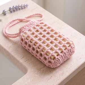 May include: A pink crocheted soap saver bag filled with soap bars, resting on a light-colored countertop. The bag has a rectangular shape with a grid-like pattern and a loop handle. A sprig of lavender is next to the bag.