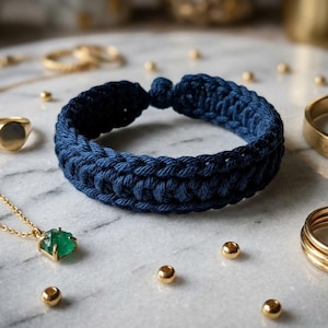 May include: A navy blue crocheted bracelet is the focal point of this image, surrounded by gold jewelry and small gold beads. A gold ring, a necklace with a green gemstone, and a gold cuff bracelet are also visible. The items are arranged on a marble surface.