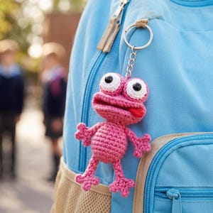 May include: A pink crocheted frog keychain with large white-eyed, hanging from a blue backpack. The frog has a red mouth and is made of a textured yarn. The backpack has a beige mesh pocket and a blue zipper.