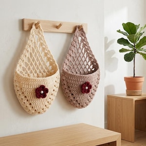 May include: Two handmade crochet hanging baskets in cream and mauve, each adorned with a burgundy flower. They hang from a wooden rack on a white wall. A small potted plant sits on a wooden side table.