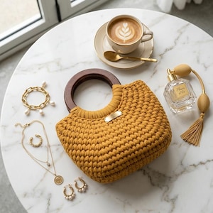 Puede incluir: Un bolso de ganchillo color mostaza con asas circulares de madera oscura y una pequeña placa rectangular dorada, acompañado de una pulsera de cadena dorada con perlas, collares dorados superpuestos, pendientes de aro, un perfume y una taza de café.