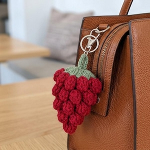 May include: A red crocheted grape cluster keychain with green leaves hangs from a brown leather handbag. The keychain is attached to a silver-colored metal clasp. The handbag has a zipper and a handle.
