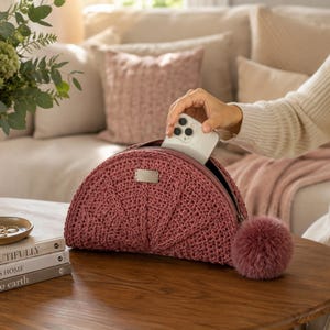 May include: A semi-circular mauve crochet pouch featuring a textured stitch pattern, a silver-toned rectangular metal tag on the front, and a matching mauve pom-pom attached to the side zip pull. A white smartphone is partially inside the pouch.