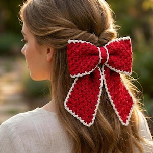 May include: A large, textured red crochet hair bow with a white border trim. The bow features a classic shape with two loops and two hanging tails, crafted with a visible yarn stitch pattern, secured to the back of long, light brown hair.