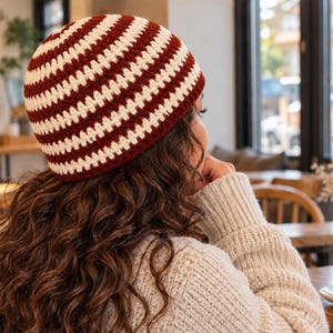 May include: A crochet beanie featuring alternating horizontal stripes in deep maroon and off-white. The hat has a rounded crown and a snug fit, constructed with a consistent stitch pattern that creates a textured surface across the entire piece.