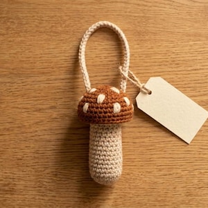 May include: A handmade crochet mushroom ornament with a brown cap and white spots, a beige stem, and a loop for hanging. A blank tag is attached with twine. The mushroom is set on a wooden surface.
