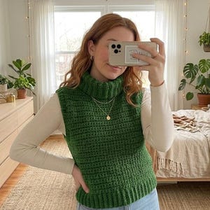 May include: A green, hand-crocheted turtleneck vest. The vest is worn over a cream-coloured long-sleeved shirt. The person is holding a phone in front of their face, and the background includes a bedroom with a bed, plants, and artwork.