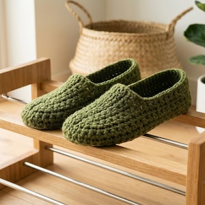 Op de afbeelding: Een paar olijfgroene gehaakte slippers met een gestructureerd ribbelpatroon. De slippers rusten op een lichthouten schoenenrek dat is voorzien van twee horizontale metalen stangen voor ondersteuning en een stevige frameconstructie.