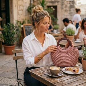 May include: A dusty rose crochet handbag featuring a rounded silhouette and a dark brown circular wooden handle. The bag has a textured, woven exterior and is held by a person wearing a white button-down shirt and gold bracelets.
