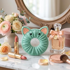 May include: A mint green crocheted cat-shaped ring with small pink inner ears and black bead eyes. The item is positioned on a white surface alongside a glass perfume bottle with a peach ribbon, two lipsticks, a makeup compact, and flowers.