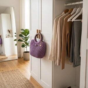 May include: A lavender crochet handbag with dark wooden circular handles hanging on a white wardrobe door. The bag features a textured knit pattern, a small rectangular brand tag, and a gold-toned circular charm attached to one of the handles.