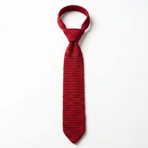 May include: A solid deep red necktie featuring a distinct knitted texture throughout the entire length of the fabric. The tie is tied in a standard knot at the top, creating a circular neck loop, and tapers down to a pointed end at the bottom.