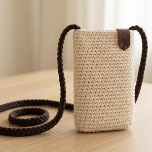 Könnte beinhalten: Eine beige gehäkelte Handytasche mit einem braunen geflochtenen Riemen. Die Tasche hat eine rechteckige Form mit einem braunen Lederakzent oben. Der Riemen ist lang genug, um sie quer über dem Körper zu tragen.