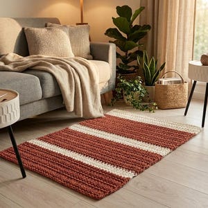 Puede incluir: Una alfombra rectangular tejida a mano con rayas alternas de color rojo óxido y crema. La alfombra está colocada sobre un suelo de madera clara en un salón, cerca de un sofá gris y una mesa auxiliar.